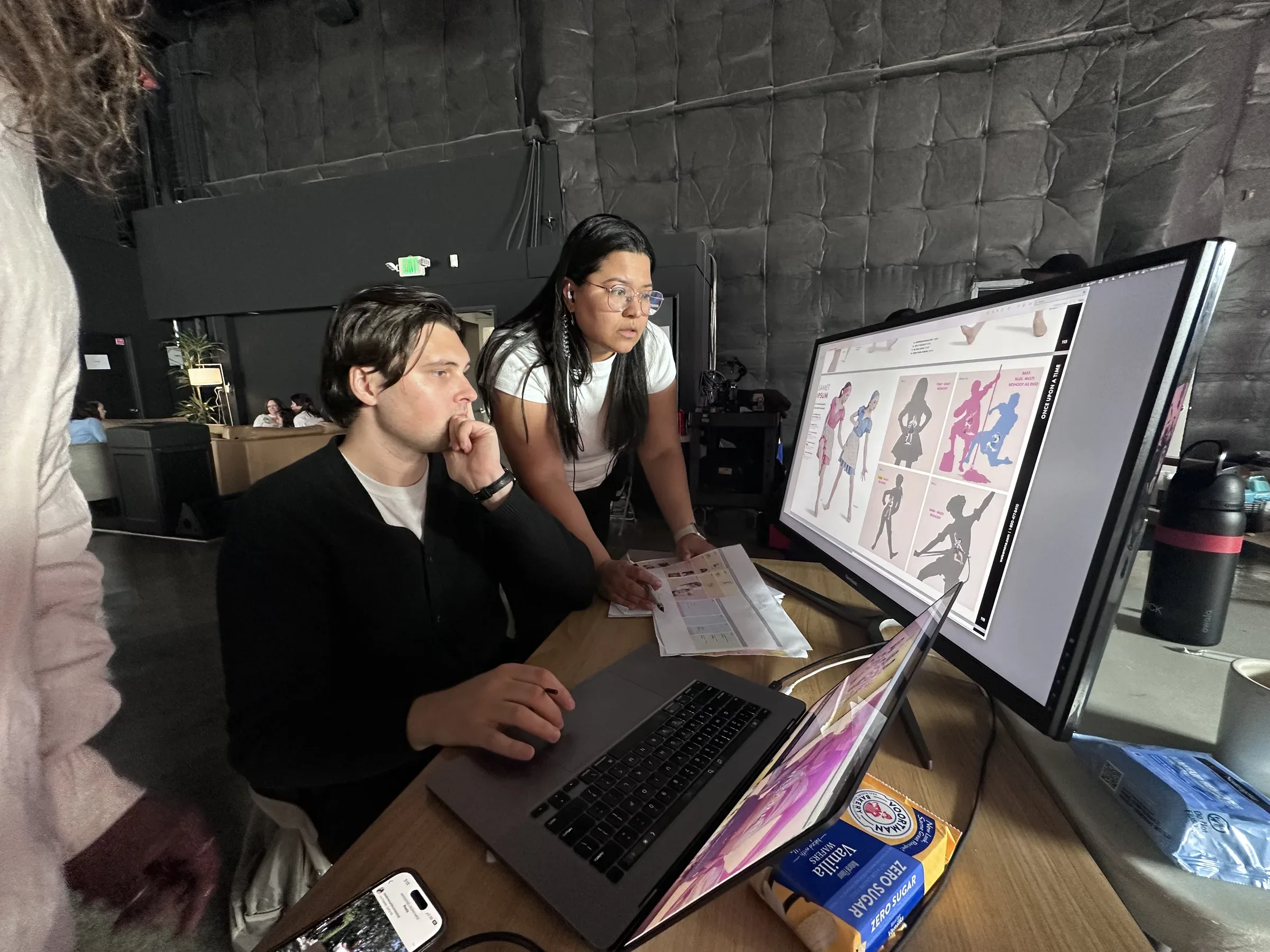 Production artist studying the monitor, chin on hand, thinking, catalog with silhouette placeholders visible on screen